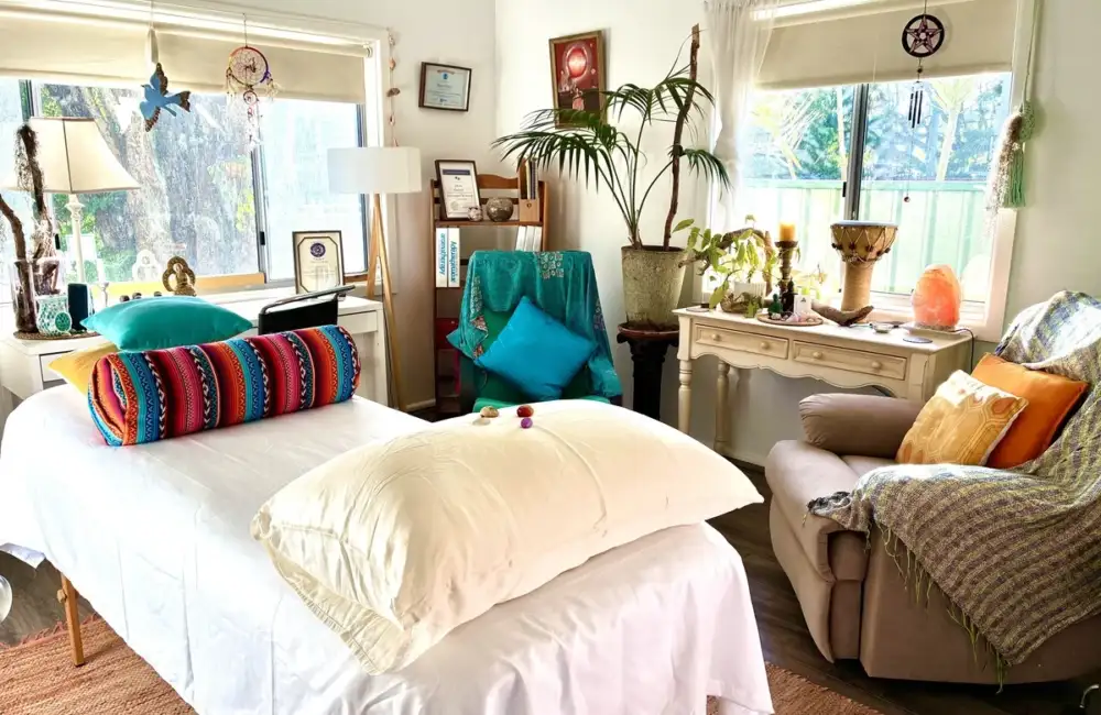 Bright bohemian therapy room with a massage table, white sheets, and a colorful bolster in front of sunlit windows.